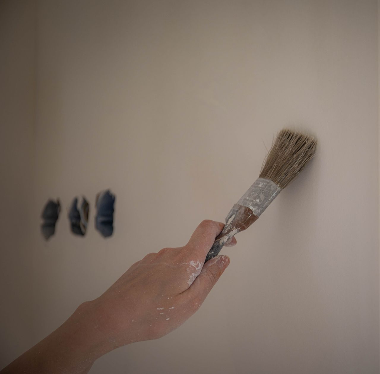 A close-up of a hand using a brush to paint a white wall, three color swatches visible.